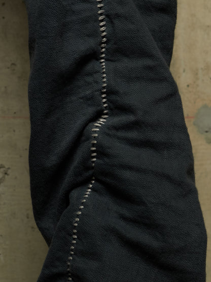Close-up of dark indigo denim fabric with prominent contrast white stitching along a seam, set against a textured concrete background.