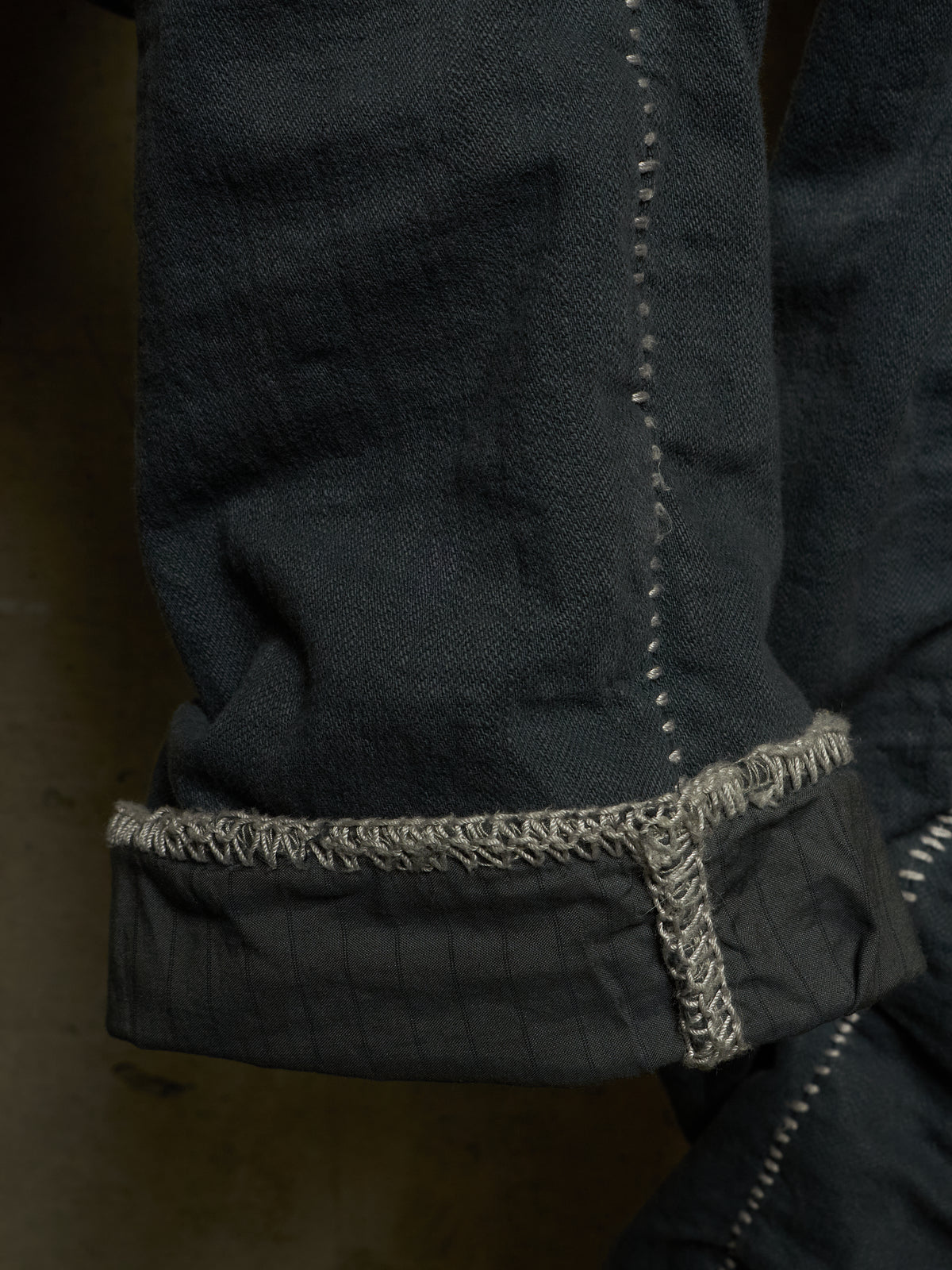 Close-up detail of a dark charcoal gray denim garment, showing a rolled cuff with contrasting light gray frayed stitching.