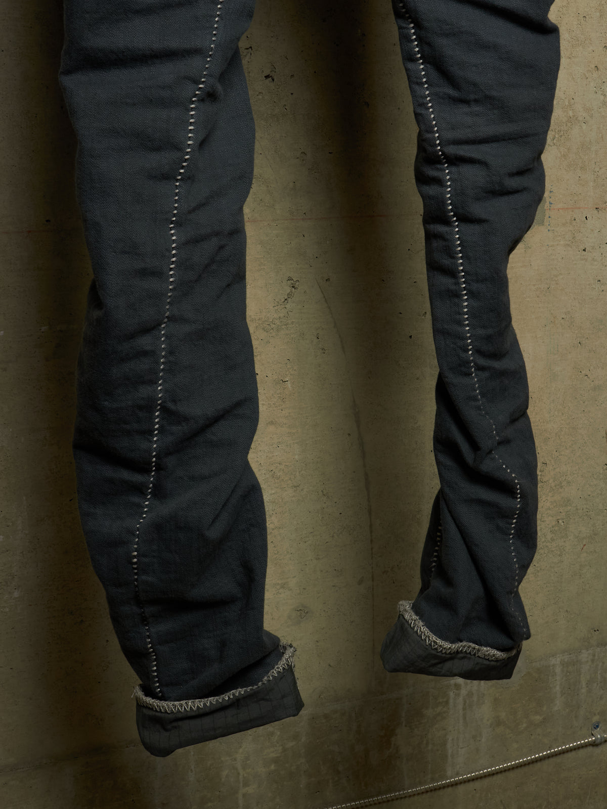 Close-up of dark charcoal gray denim jeans with rolled cuffs, showing detailed contrast stitching and a textured fabric surface against a concrete wall.