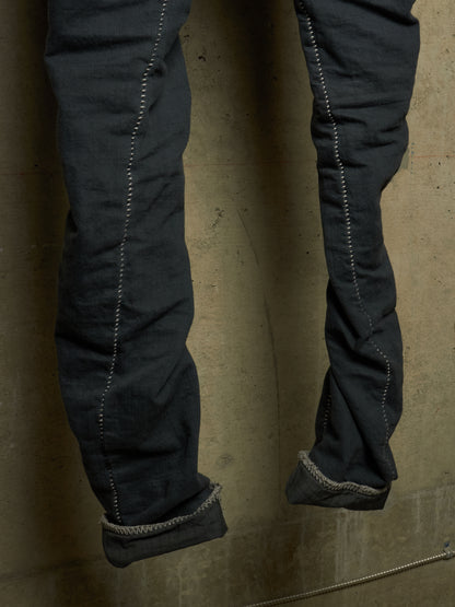 Close-up of dark charcoal gray denim jeans with rolled cuffs, showing detailed contrast stitching and a textured fabric surface against a concrete wall.