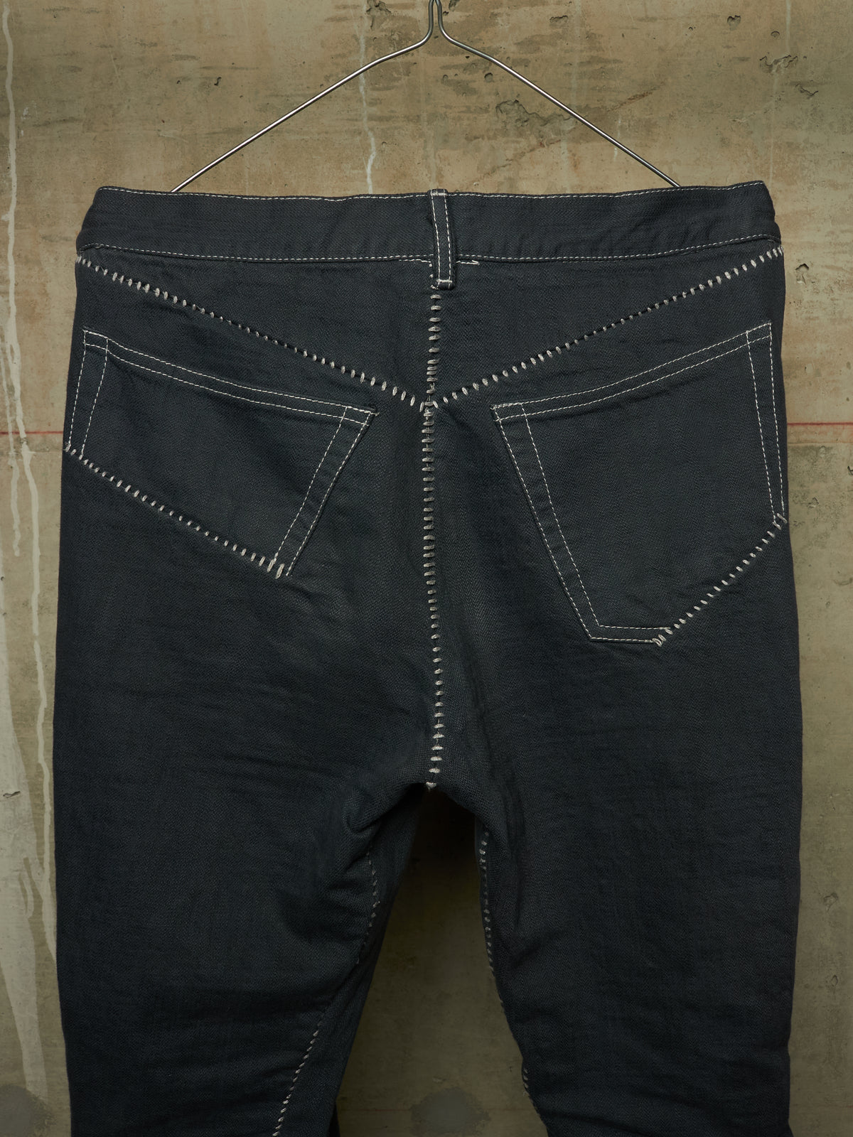 Dark charcoal gray denim jeans shown from the back, hanging on a metal hanger against a textured concrete wall. The jeans feature prominent white contrast stitching outlining the back pockets and down the center back seam.