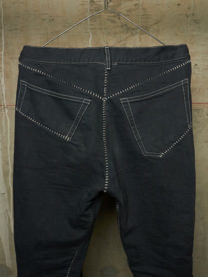 Dark charcoal gray denim jeans shown from the back, hanging on a metal hanger against a textured concrete wall. The jeans feature prominent white contrast stitching outlining the back pockets and down the center back seam.
