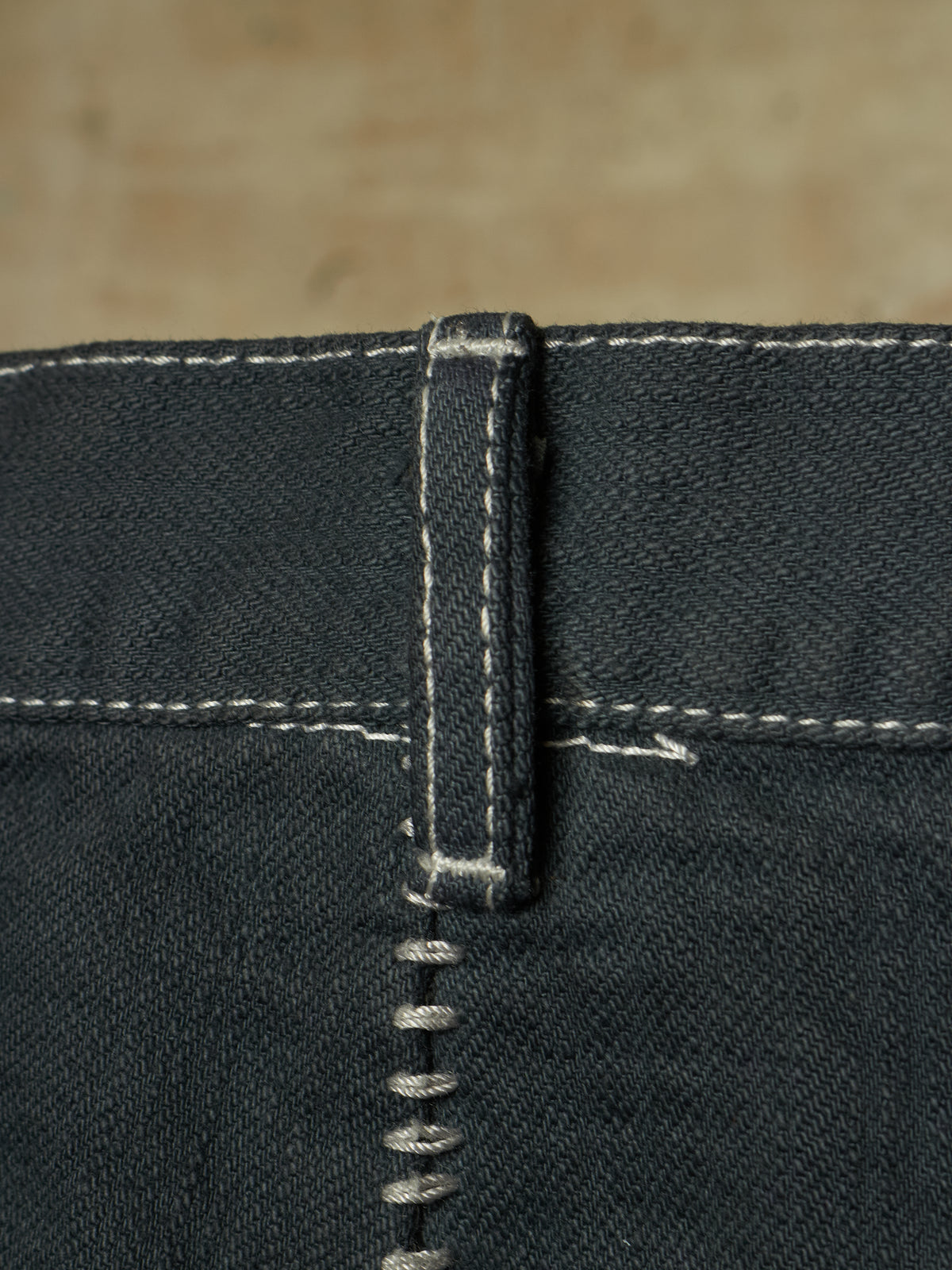 Close-up detail of dark indigo denim jeans showing the belt loop, white contrast stitching, and a silver metal rivet.