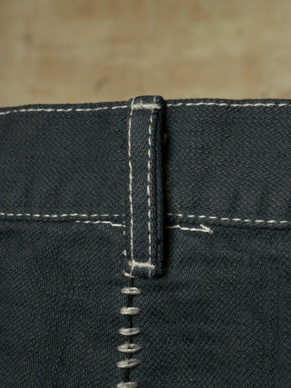 Close-up detail of dark indigo denim jeans showing the belt loop, white contrast stitching, and a silver metal rivet.
