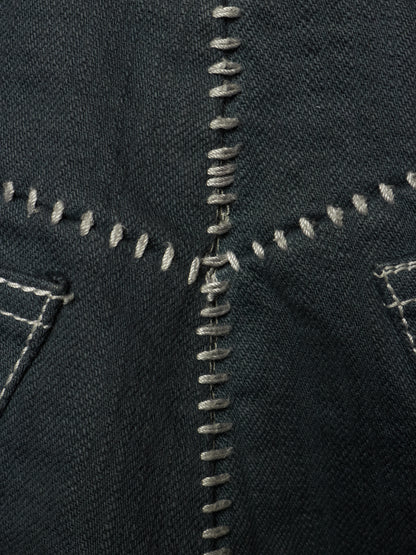 Close-up of dark indigo denim fabric with prominent contrast white and gray stitching, showing the texture of the denim weave and decorative patchwork seams.