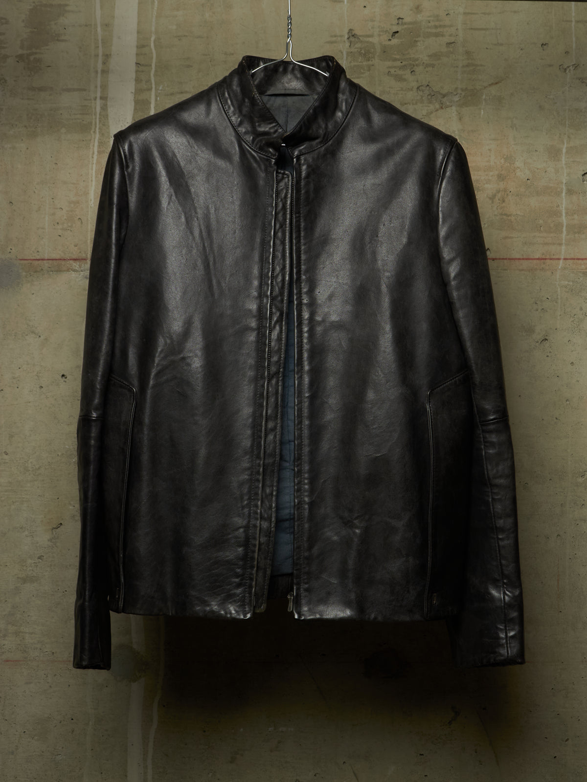 A black leather jacket with a high stand collar, hanging on a wire hanger against a textured concrete wall.