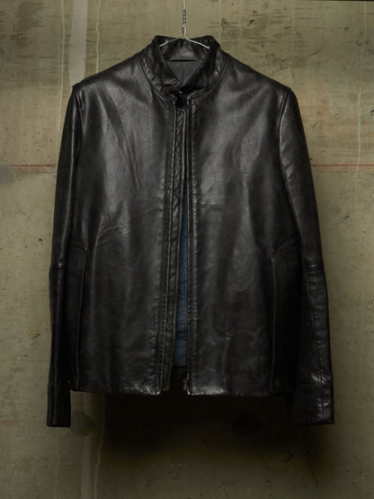 A black leather jacket with a high stand collar, hanging on a wire hanger against a textured concrete wall.