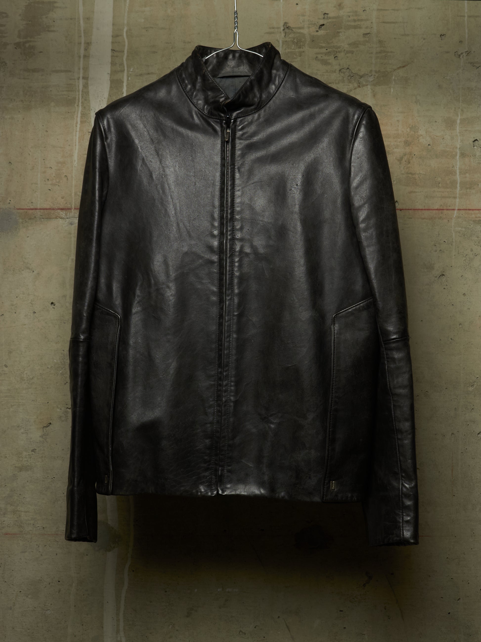 A black leather jacket with a stand collar and front zipper, hanging on a wire hanger against a textured concrete wall.
