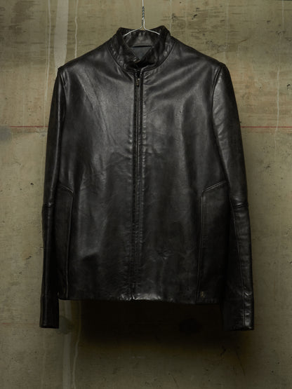 A black leather jacket with a stand collar and front zipper, hanging on a wire hanger against a textured concrete wall.