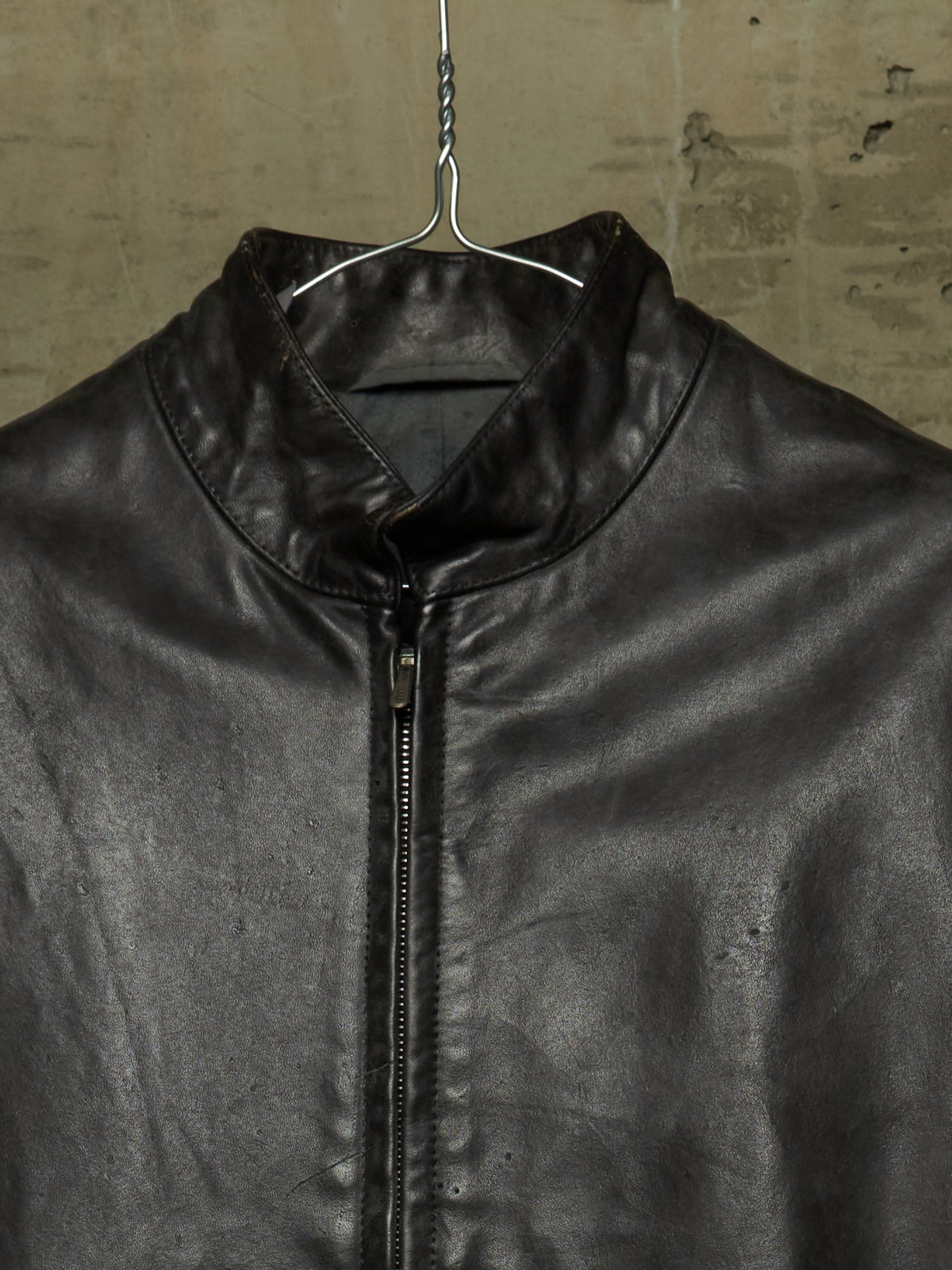 A close-up of a black leather jacket hanging on a metal hanger against a textured concrete wall. The jacket features a high collar, a front zipper, and a slightly distressed texture.