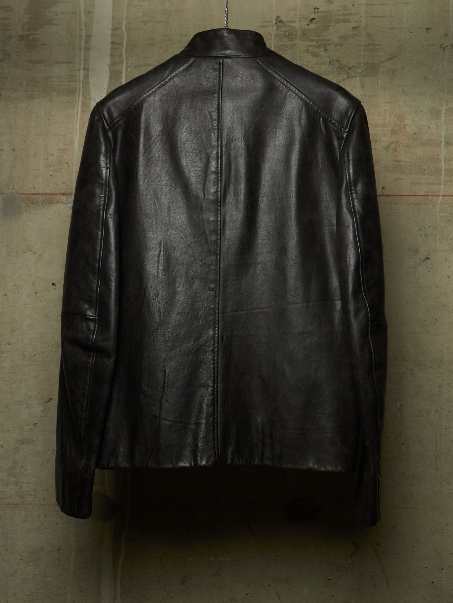 A black leather jacket shown from the back, hanging against a textured concrete wall.