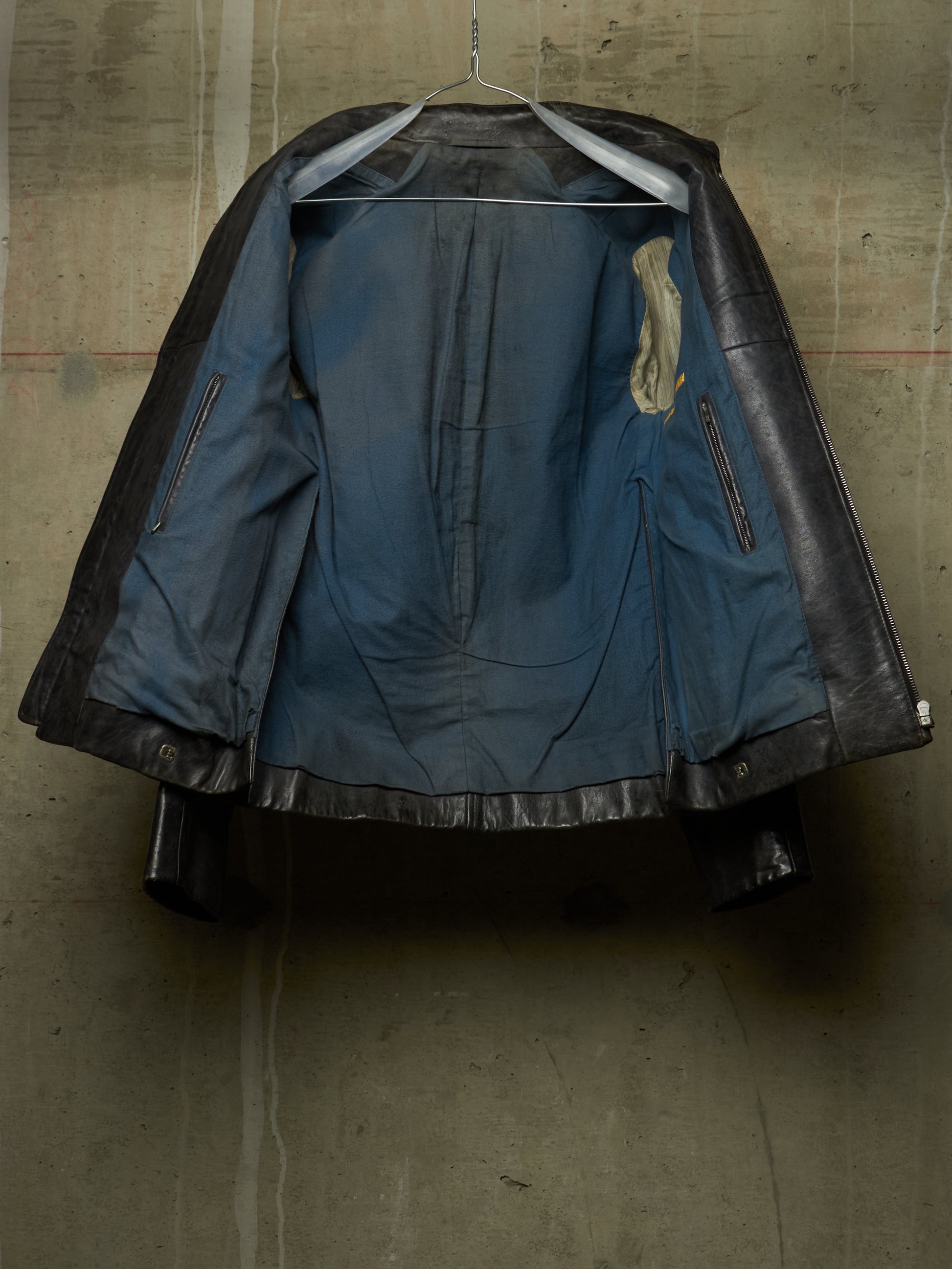 A vintage black leather jacket with a distressed blue denim lining, hanging on a wire hanger against a textured concrete wall.
