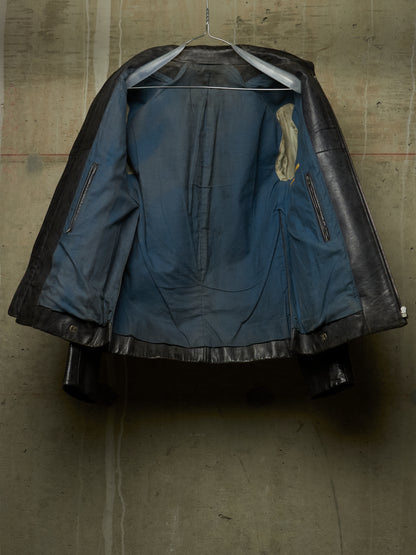 A vintage black leather jacket with a distressed blue denim lining, hanging on a wire hanger against a textured concrete wall.