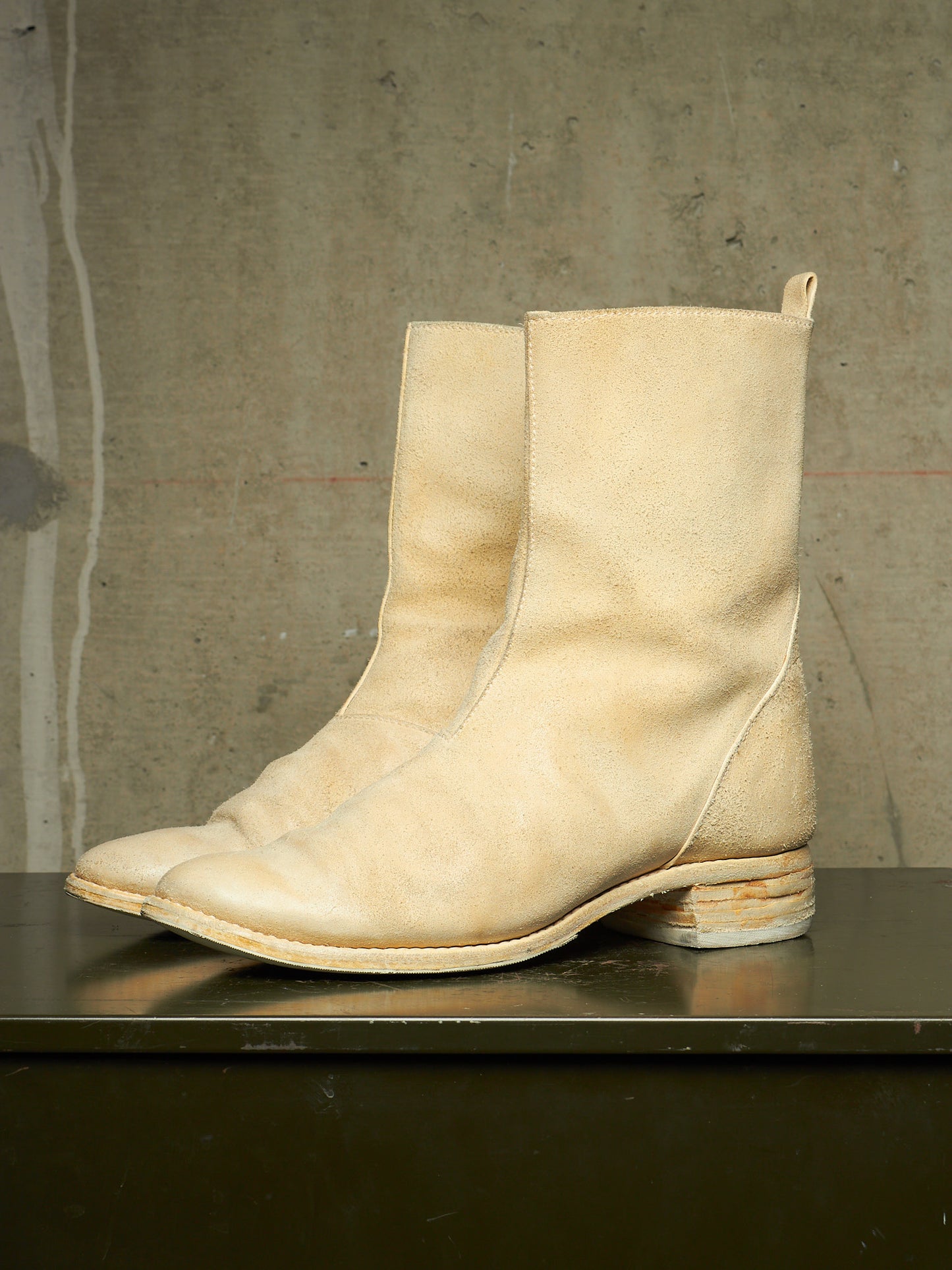 A pair of light beige leather ankle boots with a low stacked block heel, displayed on a dark surface against a textured concrete wall.