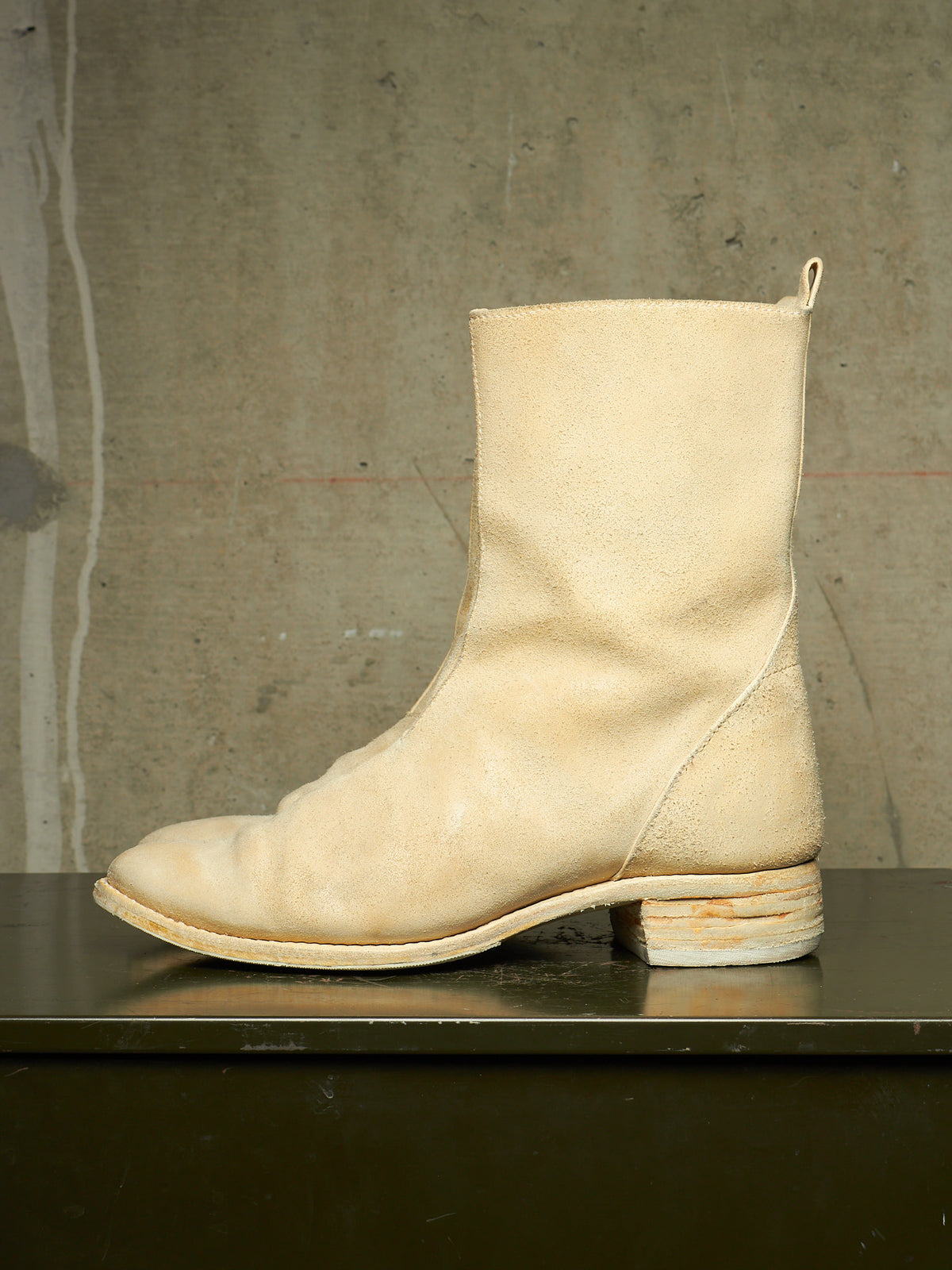 A single light beige leather ankle boot with a low stacked block heel, shown in profile on a dark surface against a textured concrete wall.