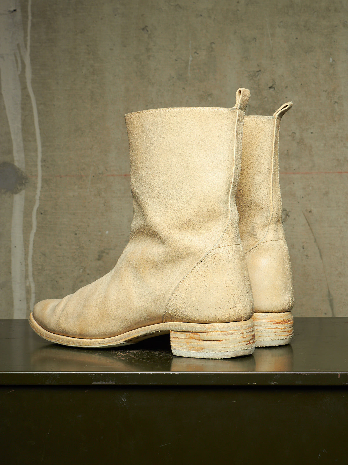 A pair of off-white leather ankle boots with stacked cork-look heels, displayed on a dark reflective surface against a textured concrete wall.