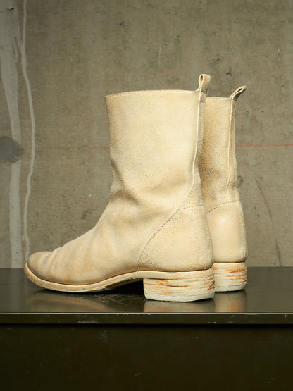 A pair of off-white leather ankle boots with stacked cork-look heels, displayed on a dark reflective surface against a textured concrete wall.