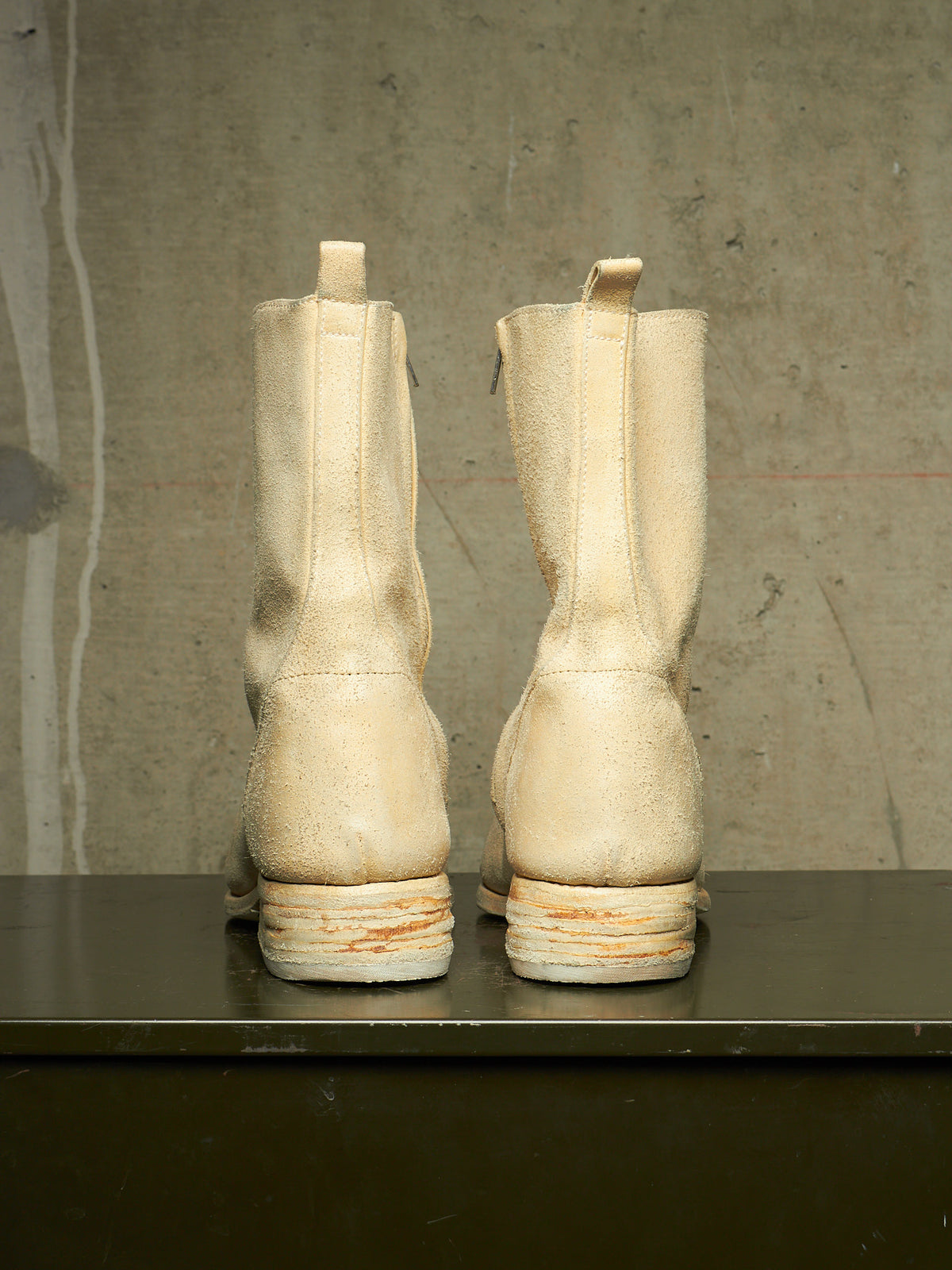A pair of off-white distressed leather mid-calf boots shown from the back, resting on a dark surface against a textured concrete wall.