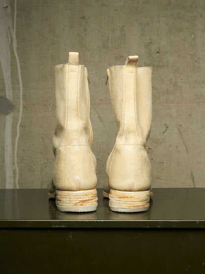 A pair of off-white distressed leather mid-calf boots shown from the back, resting on a dark surface against a textured concrete wall.