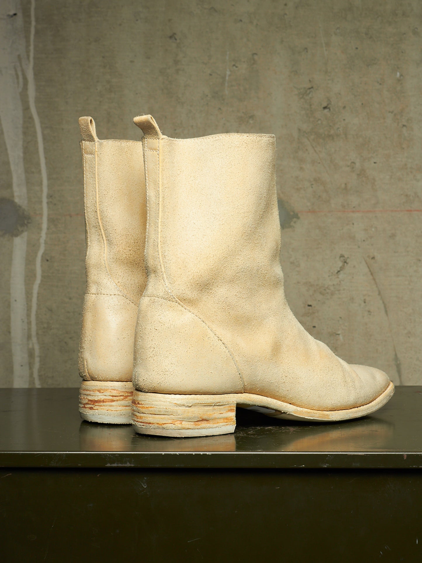 A pair of off-white suede ankle boots with stacked cork-look heels, displayed on a dark reflective surface against a textured concrete wall.