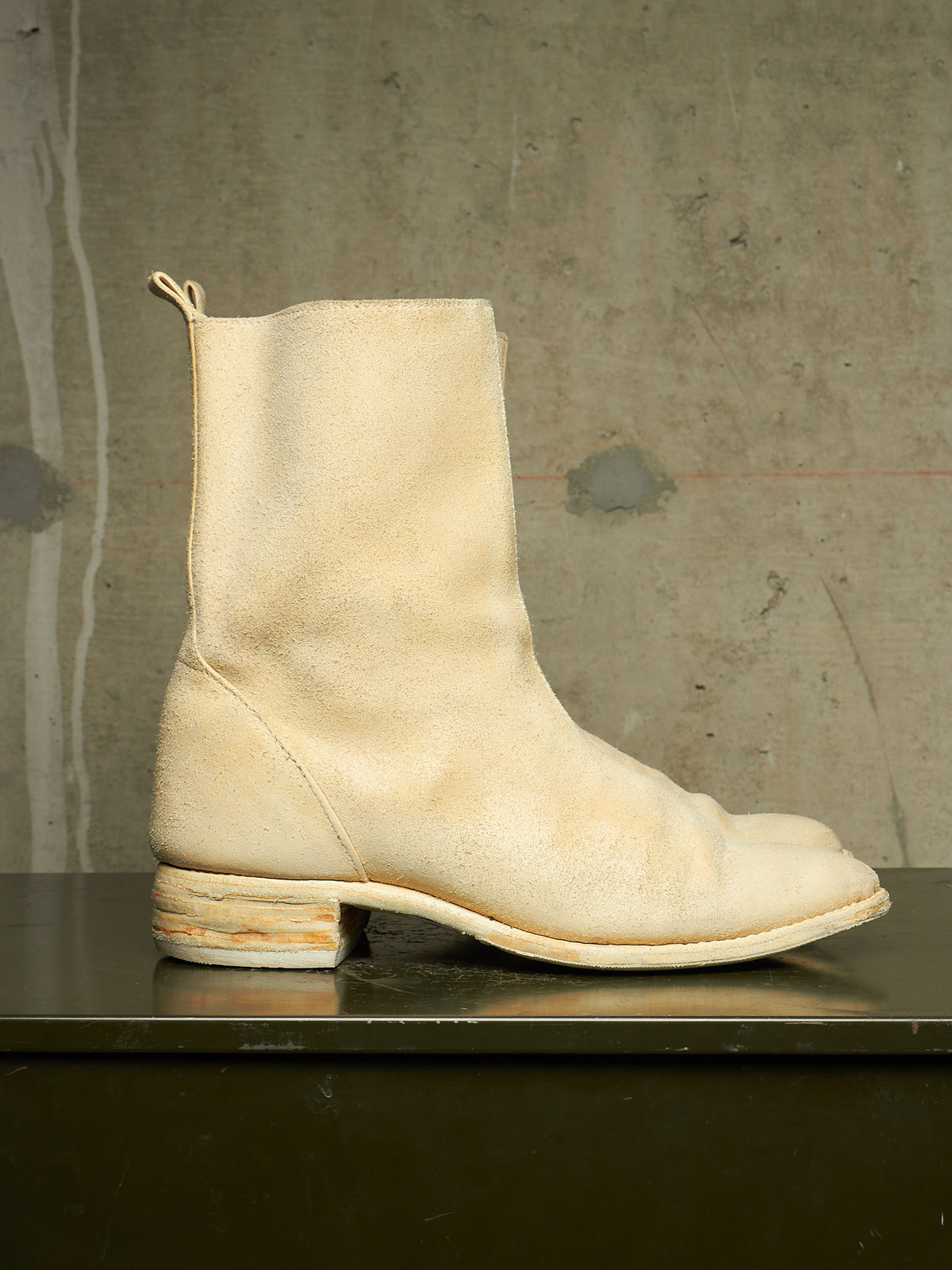 A single off-white leather ankle boot with a low stacked block heel, shown in profile on a dark reflective surface against a textured concrete wall.