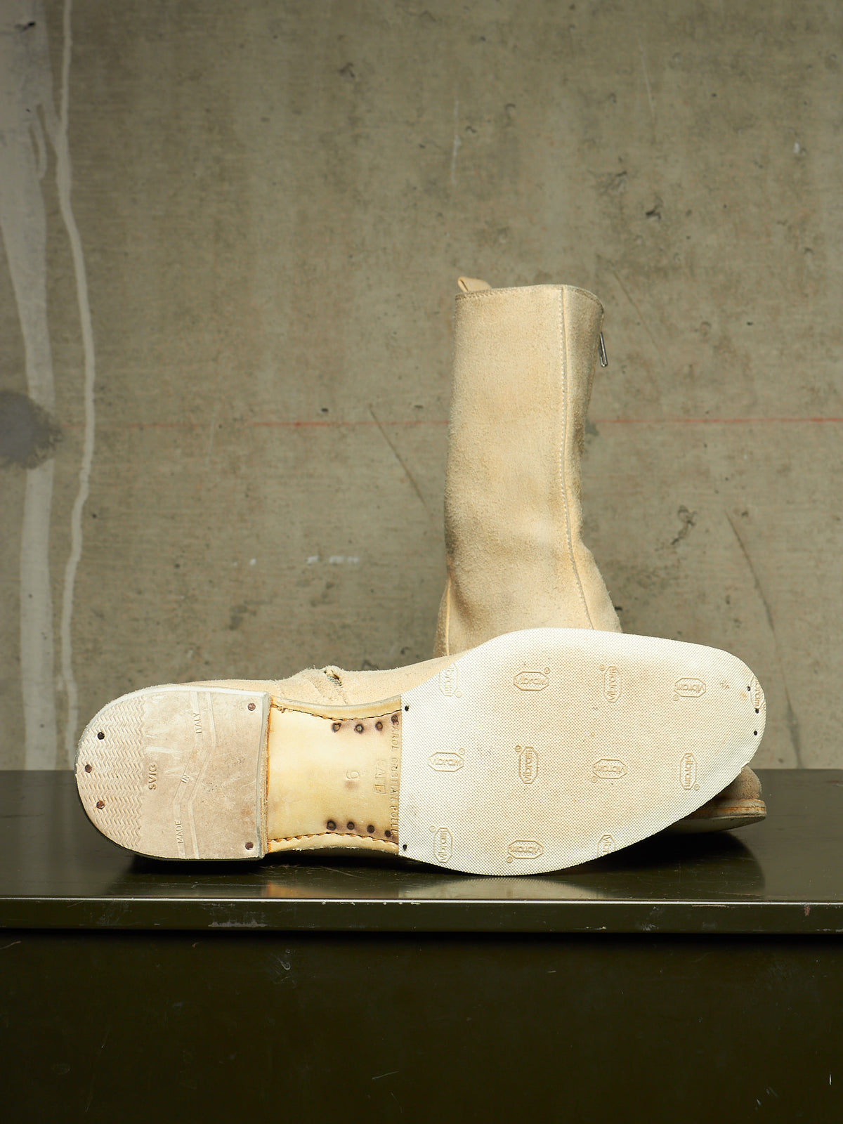 A pair of tall beige leather boots with heavily worn, scuffed soles, displayed on a dark surface against a textured concrete wall.