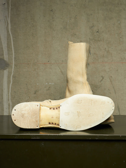 A pair of tall beige leather boots with heavily worn, scuffed soles, displayed on a dark surface against a textured concrete wall.