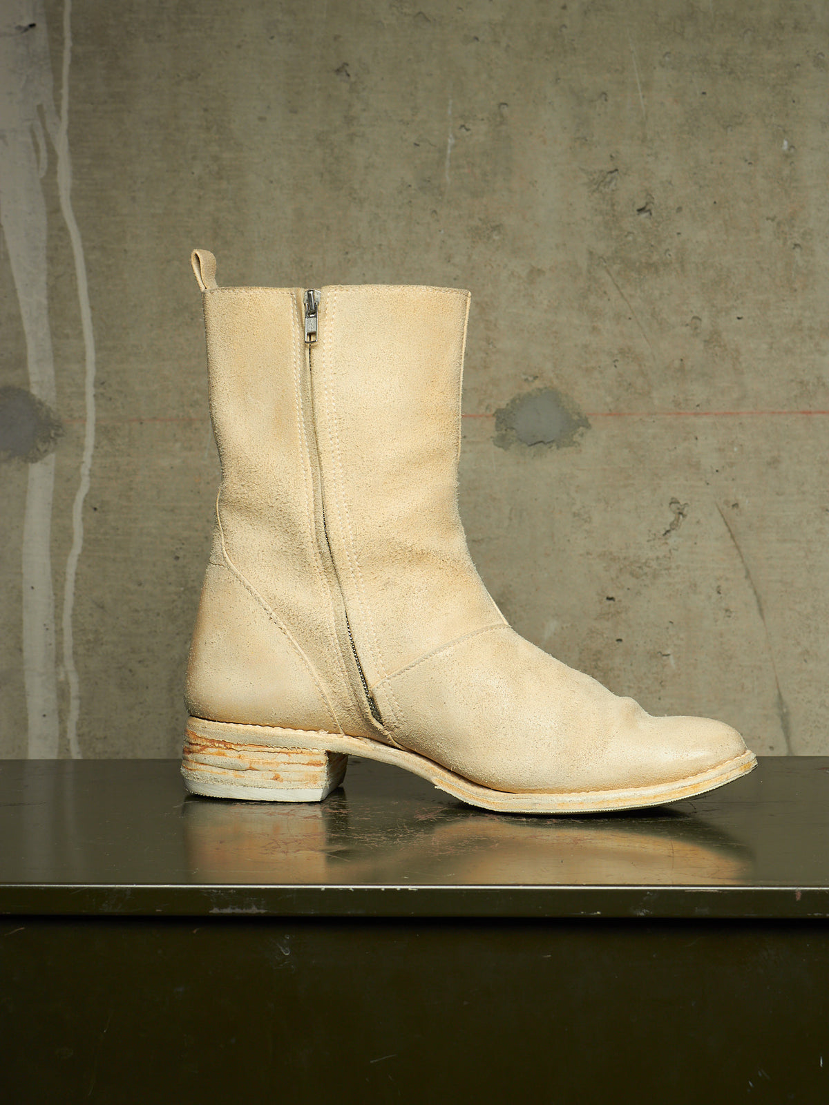 A light beige leather ankle boot with a side zipper, square toe, and stacked block heel, displayed on a dark reflective surface against a textured concrete wall.