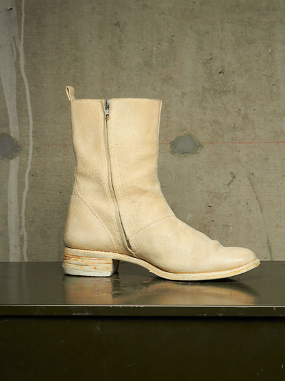 A light beige leather ankle boot with a side zipper, square toe, and stacked block heel, displayed on a dark reflective surface against a textured concrete wall.
