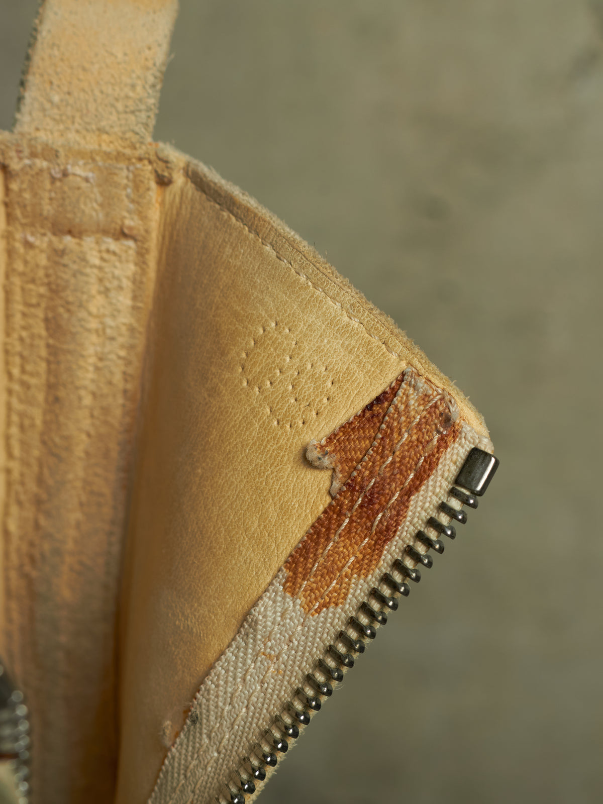 Close-up of a beige fabric accessory showing a worn, frayed section with exposed brown lining and a metal zipper along the edge, set against a neutral gray background.