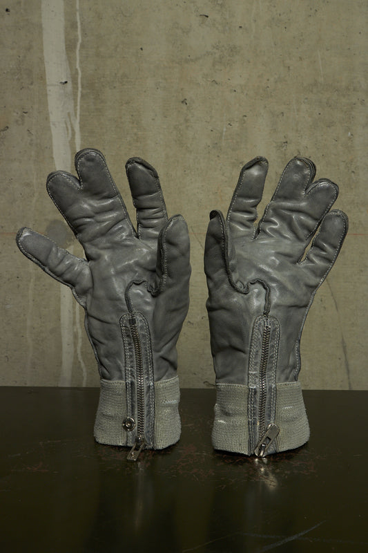 A pair of gray leather gloves with built-in zippers at the wrist, displayed on a dark reflective surface against a textured concrete wall.