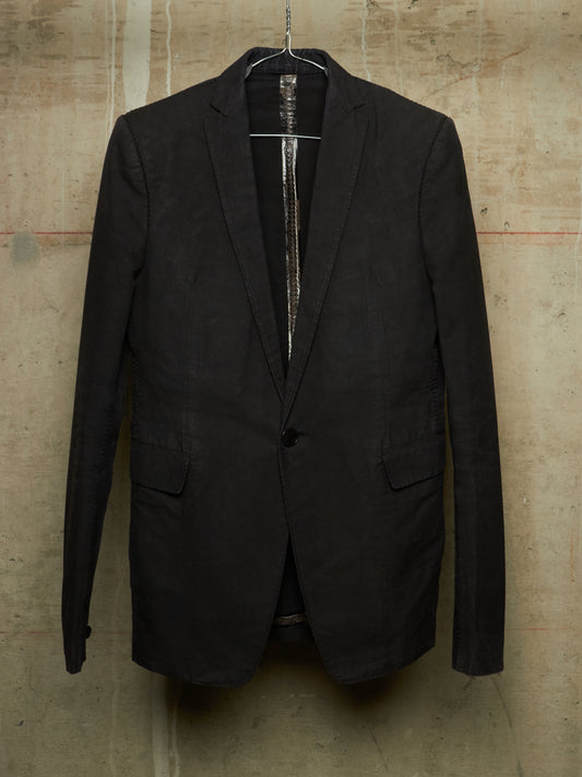 A black single-breasted blazer jacket hanging on a wire hanger against a textured concrete wall.