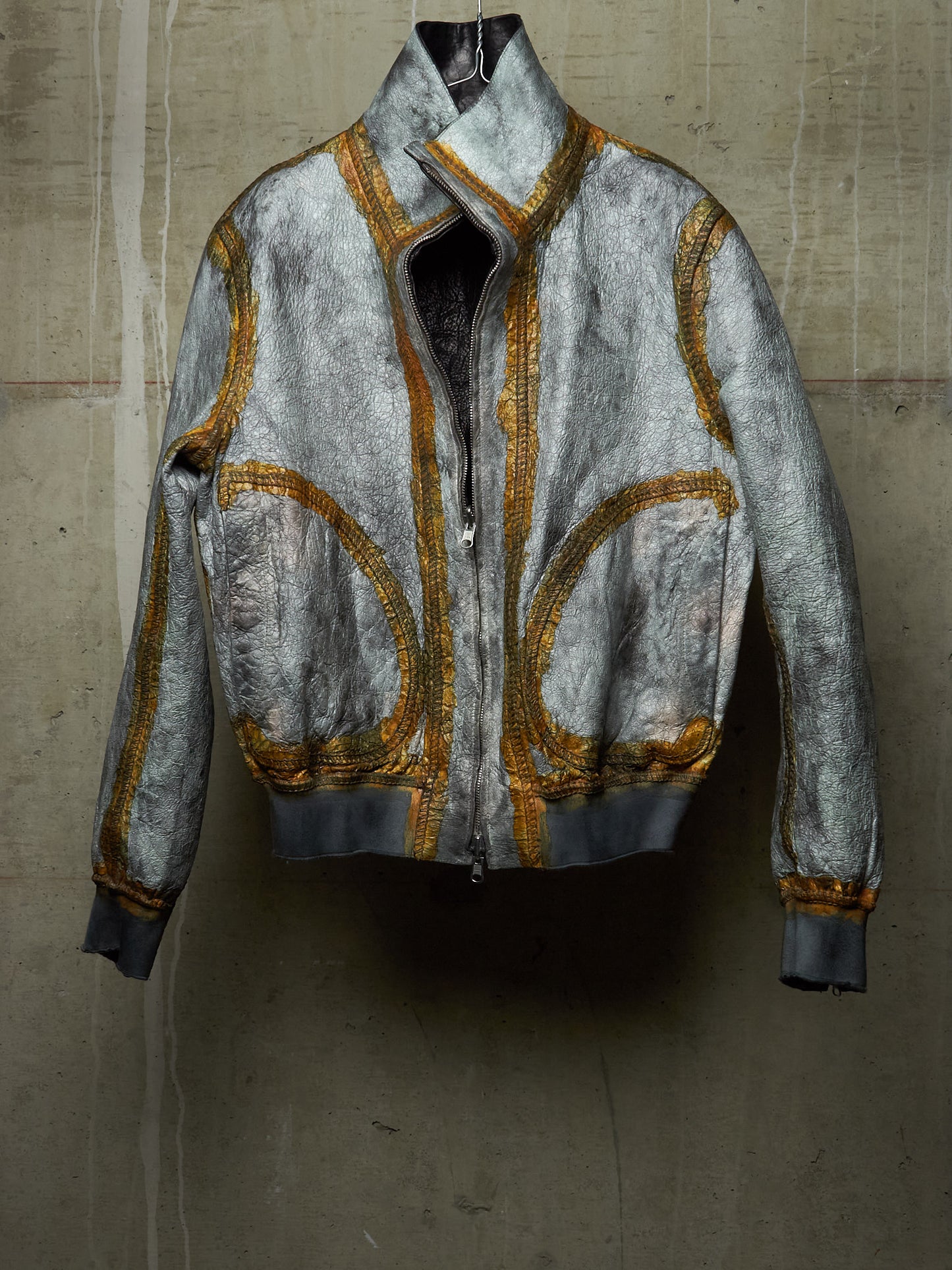 A distressed silver-gray bomber jacket with gold metallic embroidery and a high collar, displayed against a textured concrete wall.