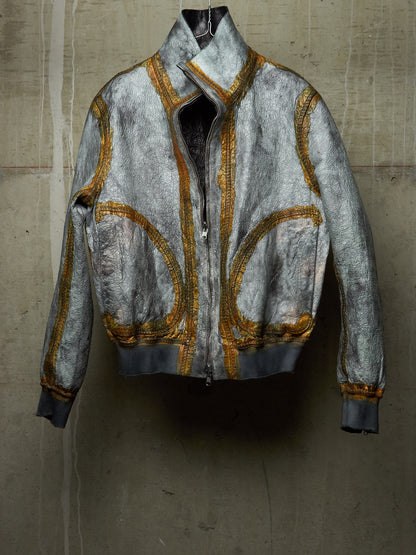 A distressed silver-gray bomber jacket with gold metallic embroidery and a high collar, displayed against a textured concrete wall.