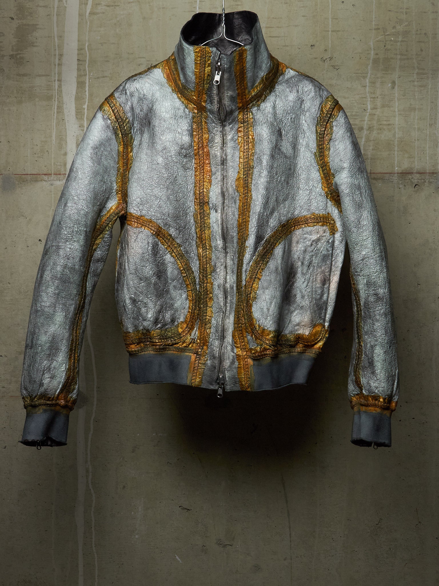 A distressed silver-gray bomber jacket with gold embroidered detailing, displayed against a textured concrete wall.