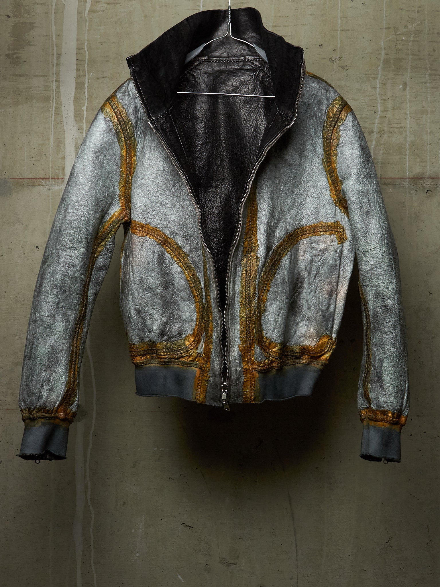 A distressed silver leather jacket with gold contrast stitching and a black interior lining, hanging against a textured concrete wall.