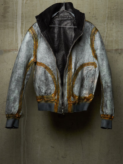 A distressed silver leather jacket with gold contrast stitching and a black interior lining, hanging against a textured concrete wall.