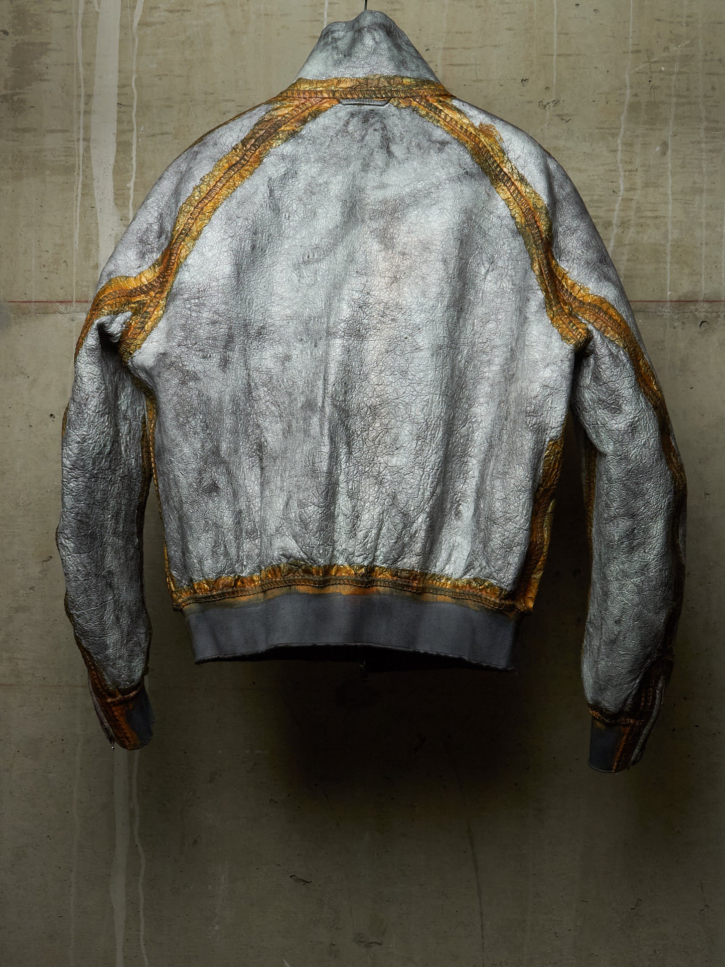 A distressed silver-gray bomber jacket with gold trim, shown from the back against a textured concrete wall.