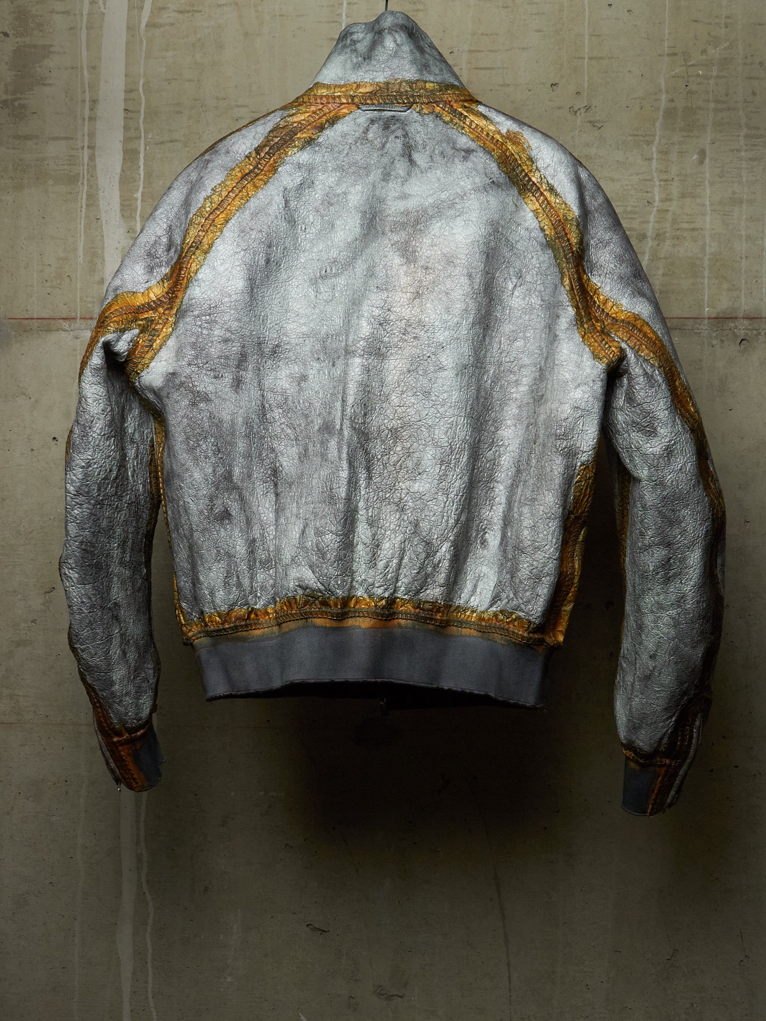 A distressed silver-gray bomber jacket with gold trim, shown from the back against a textured concrete wall.