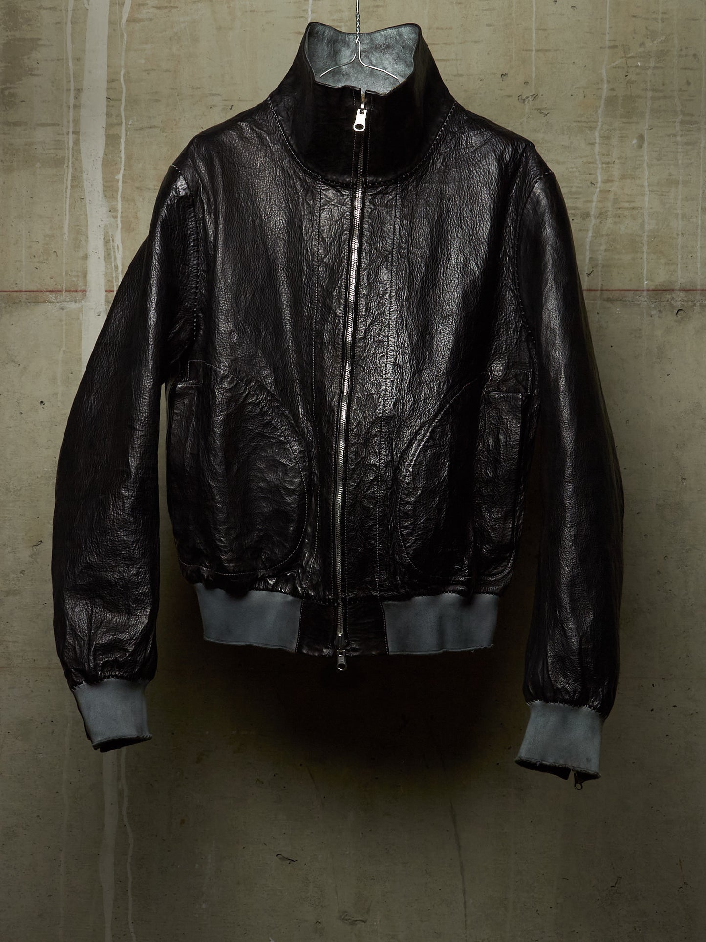A black leather bomber jacket with a high collar and silver zipper, hanging against a textured concrete wall.
