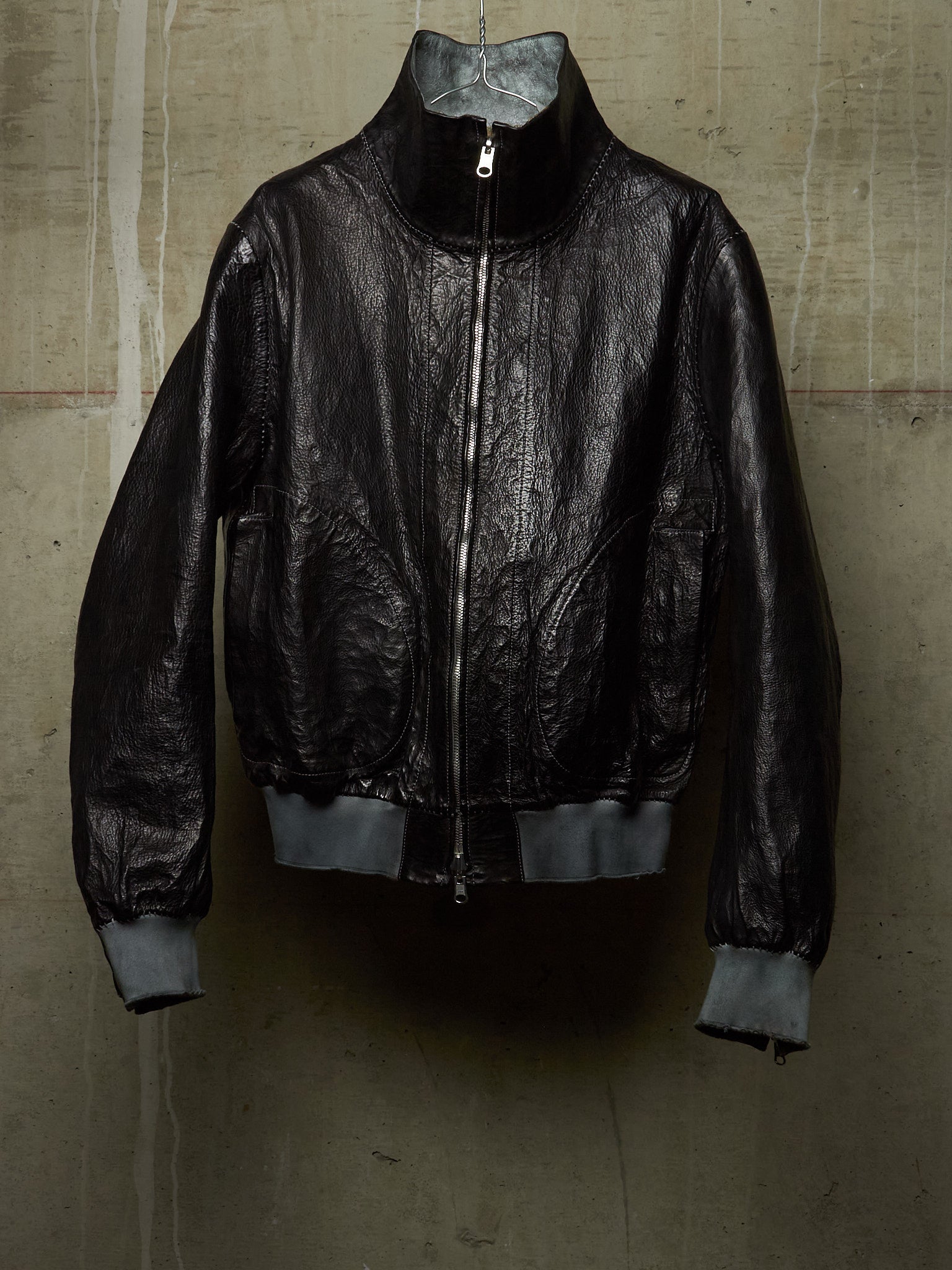 A black leather bomber jacket with a high collar and silver zipper, hanging against a textured concrete wall.