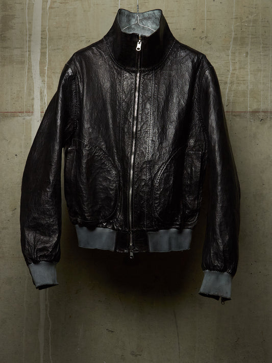A black leather bomber jacket with a high collar and silver zipper, hanging against a textured concrete wall.