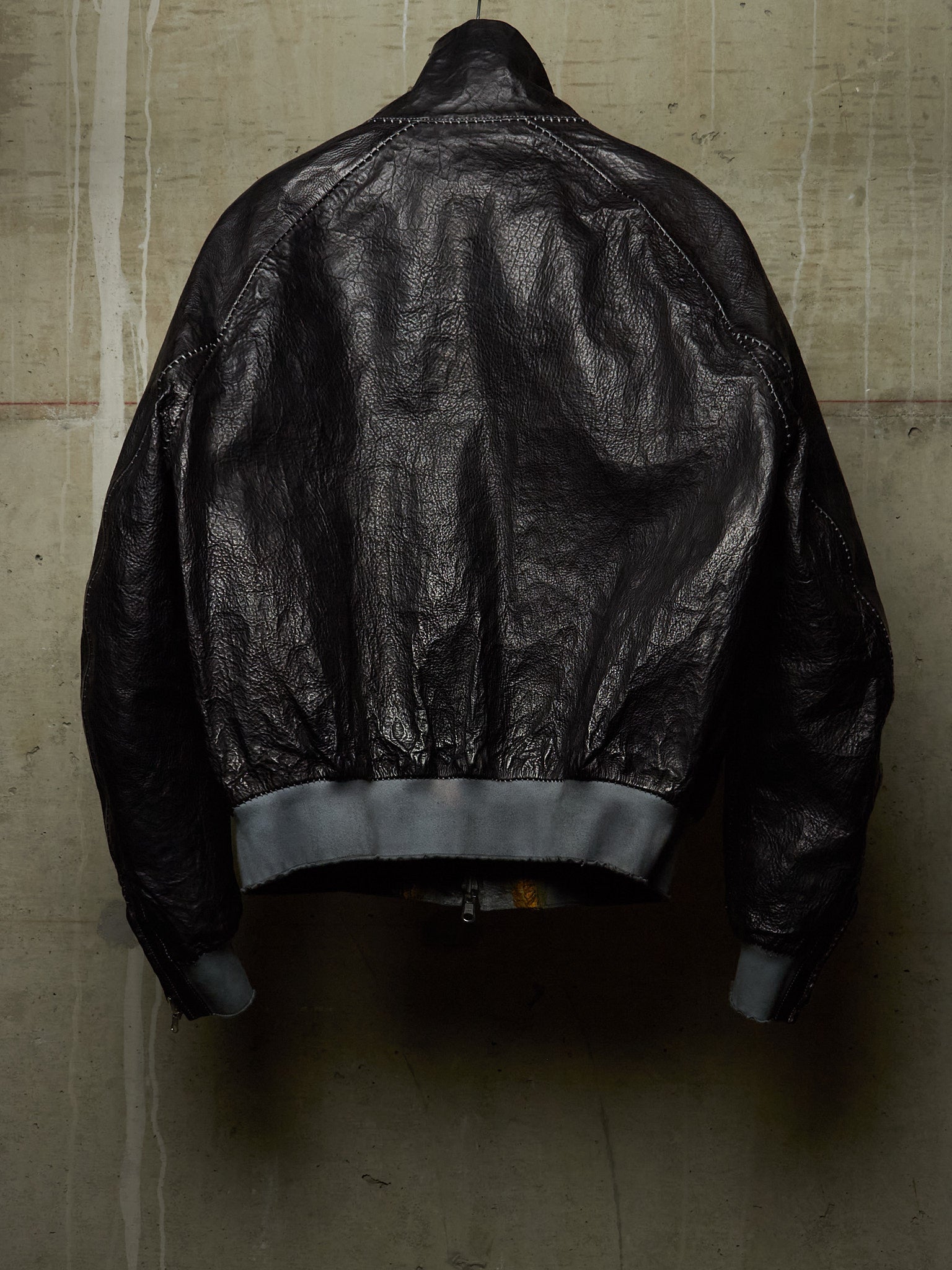 A black leather bomber jacket shown from the back, hanging against a textured concrete wall. The jacket features a high collar, zippered back, and ribbed gray cuffs and hem.