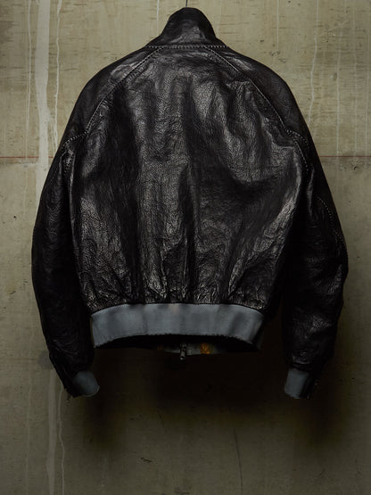 A black leather bomber jacket shown from the back, hanging against a textured concrete wall. The jacket features a high collar, zippered back, and ribbed gray cuffs and hem.