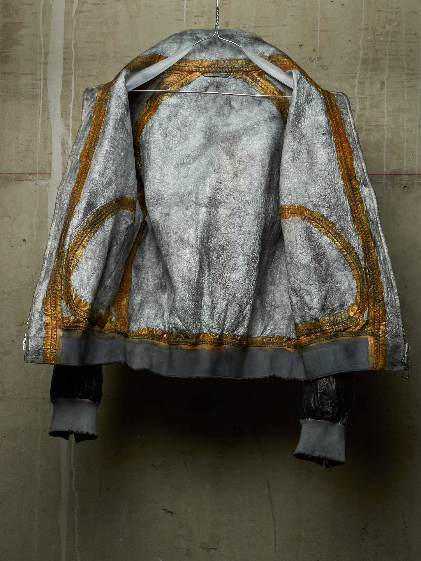 A distressed silver-gray jacket with gold embroidered circular patterns and a dark gray cuff, hanging on a wire hanger against a textured concrete wall.