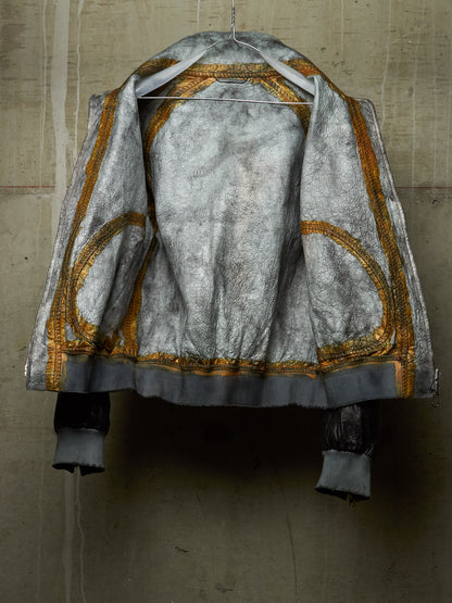 A distressed silver-gray jacket with gold embroidered circular patterns and a dark gray cuff, hanging on a wire hanger against a textured concrete wall.