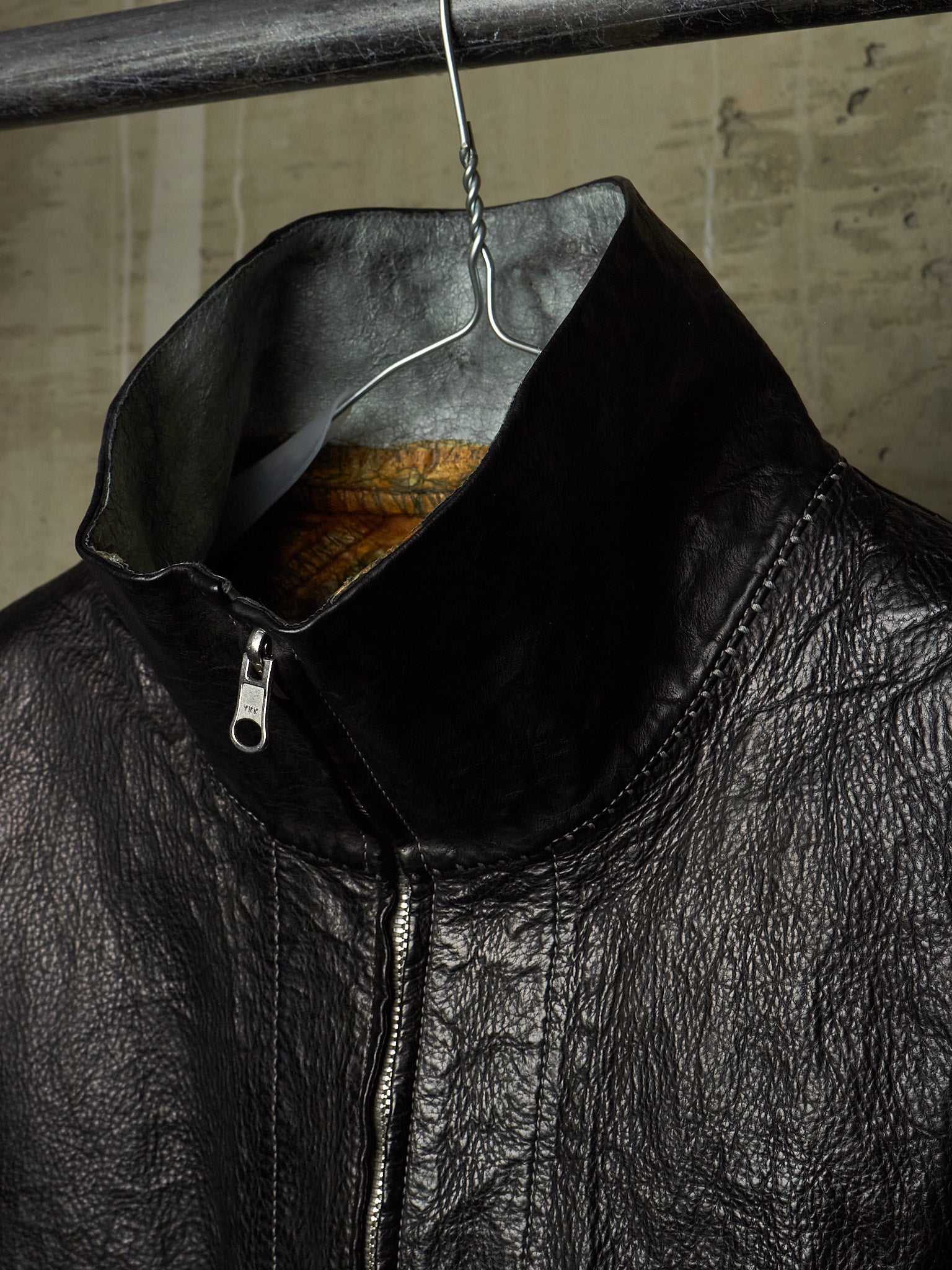 Close-up of a black leather jacket hanging on a metal hanger against a textured concrete wall. The jacket features a high collar, a silver zipper, and a textured leather surface.