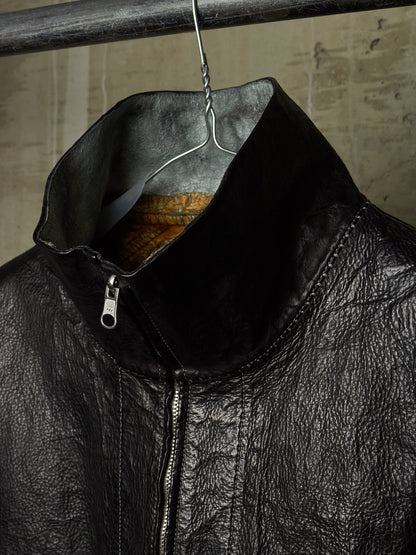 Close-up of a black leather jacket hanging on a metal hanger against a textured concrete wall. The jacket features a high collar, a silver zipper, and a textured leather surface.