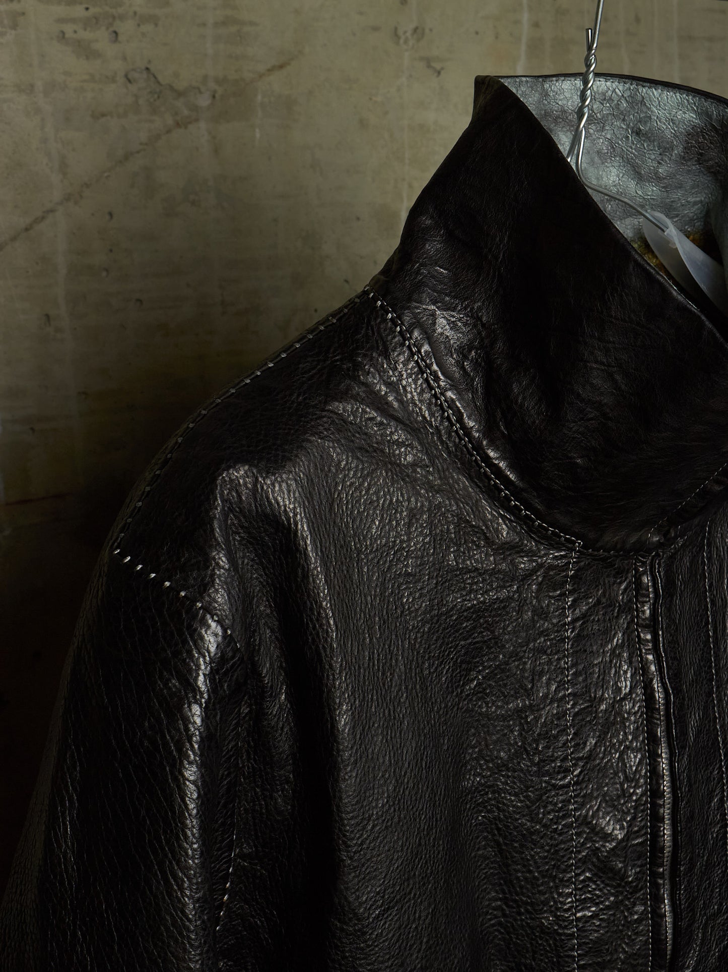 Close-up of a black leather jacket with a high collar, hanging on a metal hanger against a textured concrete wall.