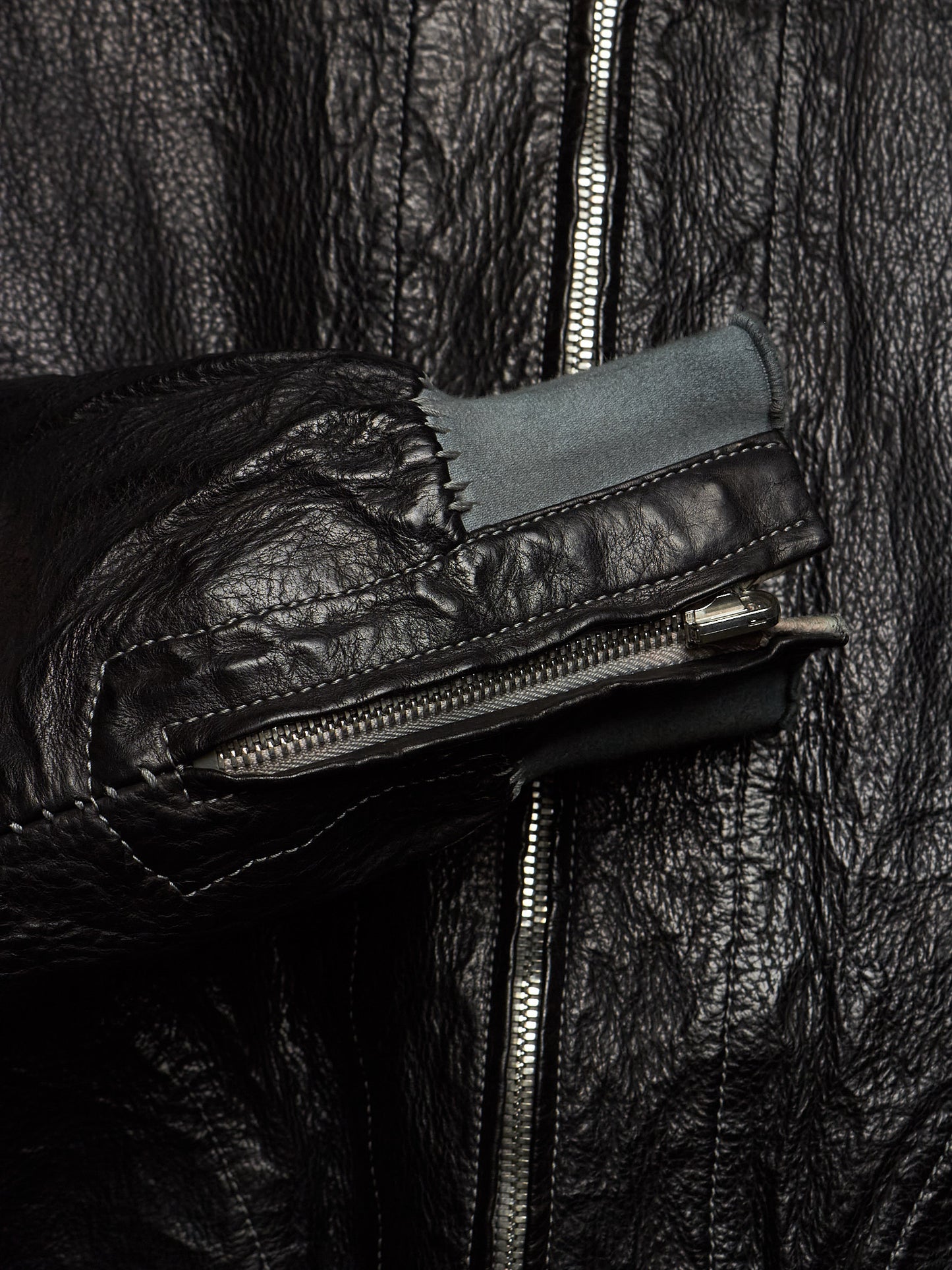 Close-up of a black leather jacket with a prominent front zipper, a zippered pocket on the sleeve, and a contrasting dark gray fabric lining.